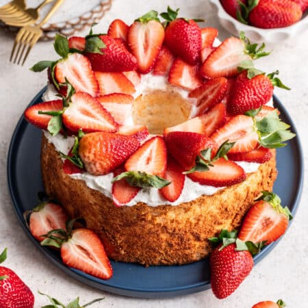 A homemade angel food cake topped with whipped cream and sliced strawberries on a blue platter.