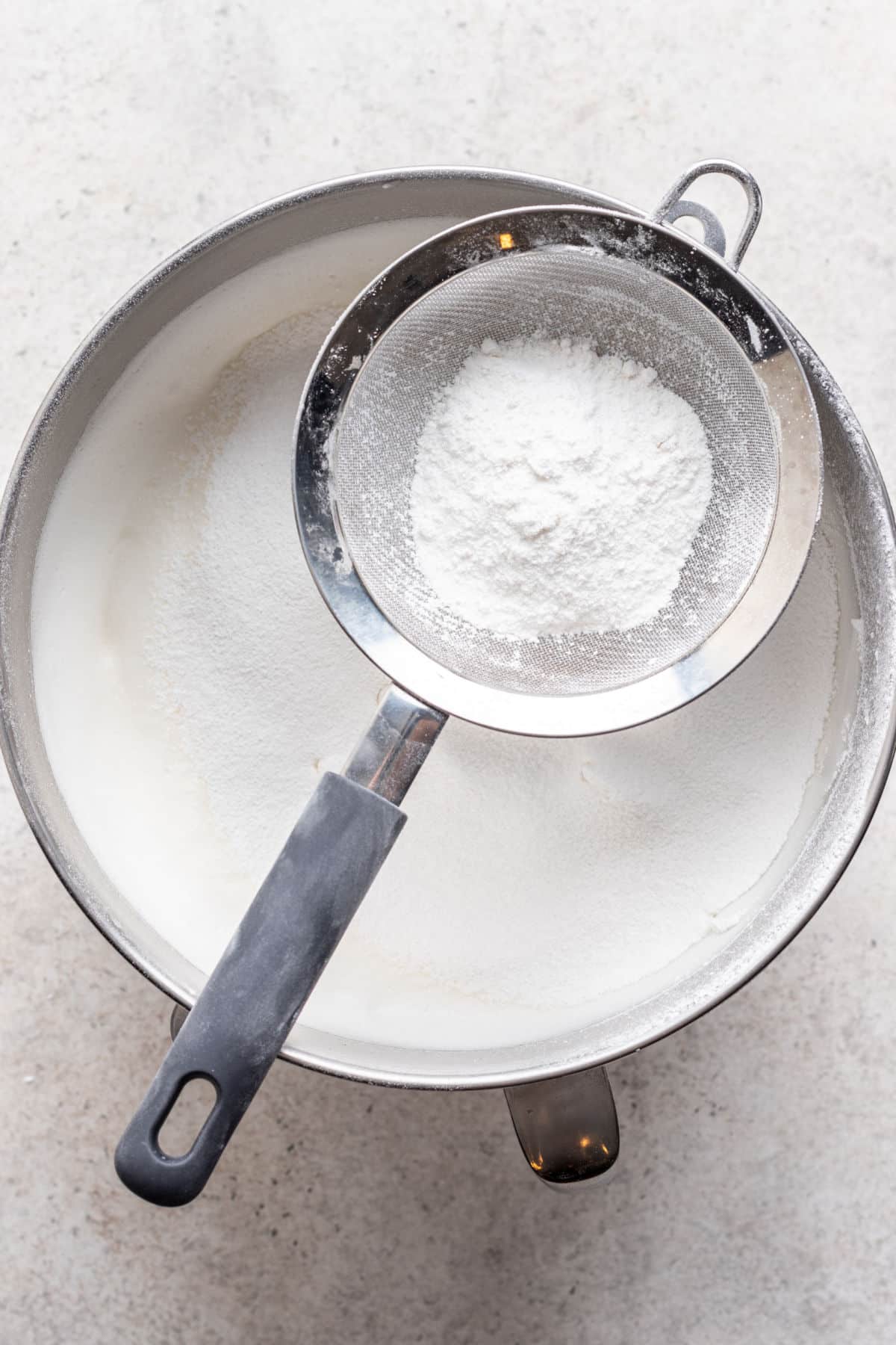 A sifter holding cake flour over a bowl of beaten egg white. 