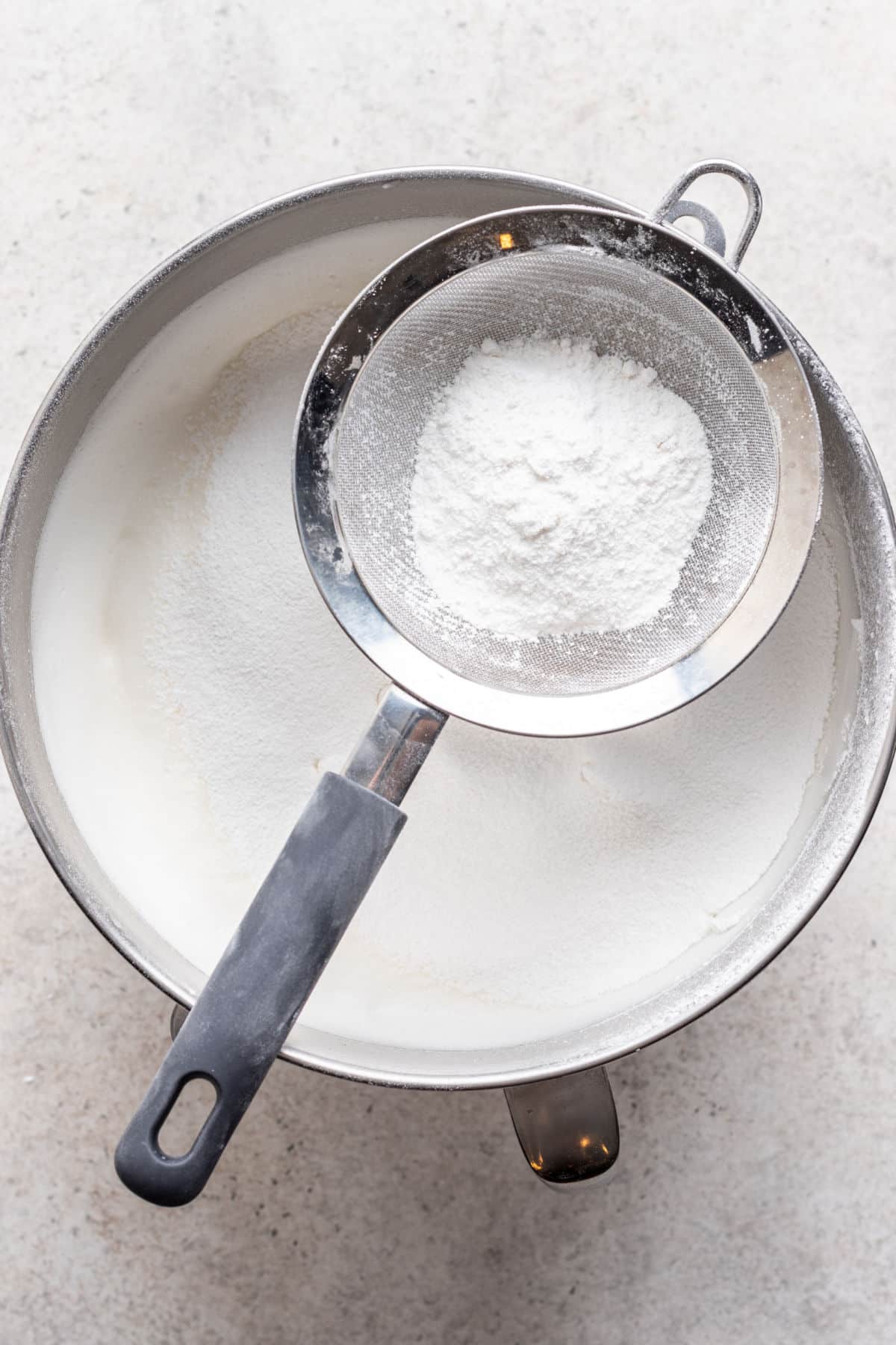 A sifter holding cake flour over a bowl of beaten egg white.