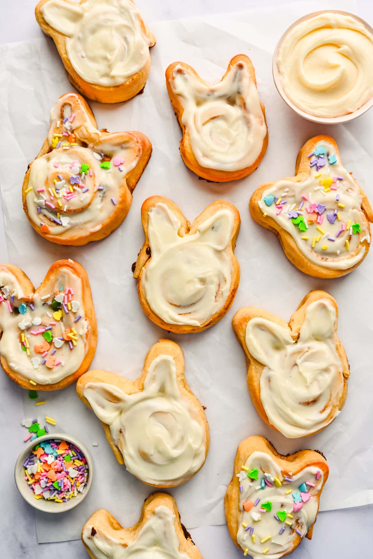 Easter bunny cinnamon rolls in a piece of parchment paper next to dishes of sprinkles and frosting.