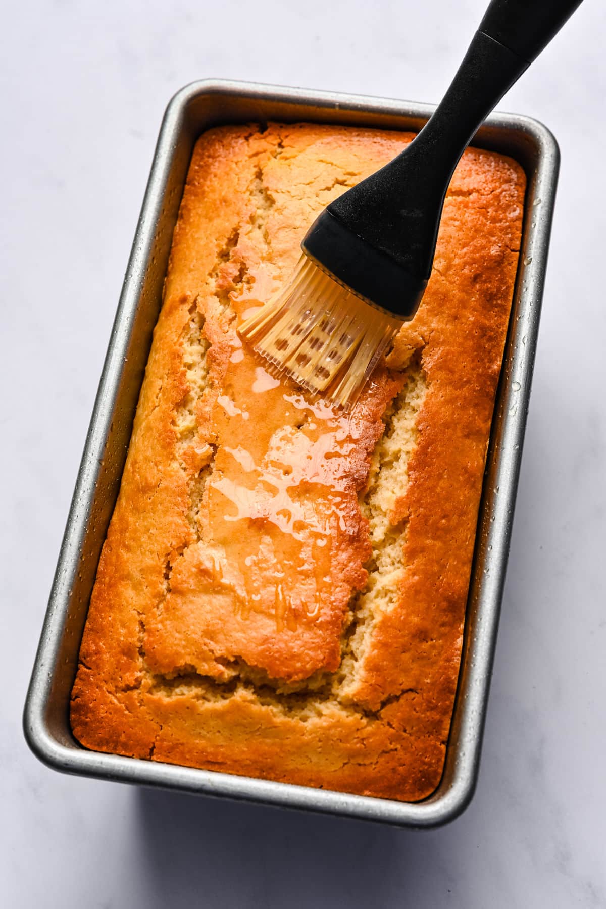 A basting brush brushing lemon syrup on top of lemon loaf cake.