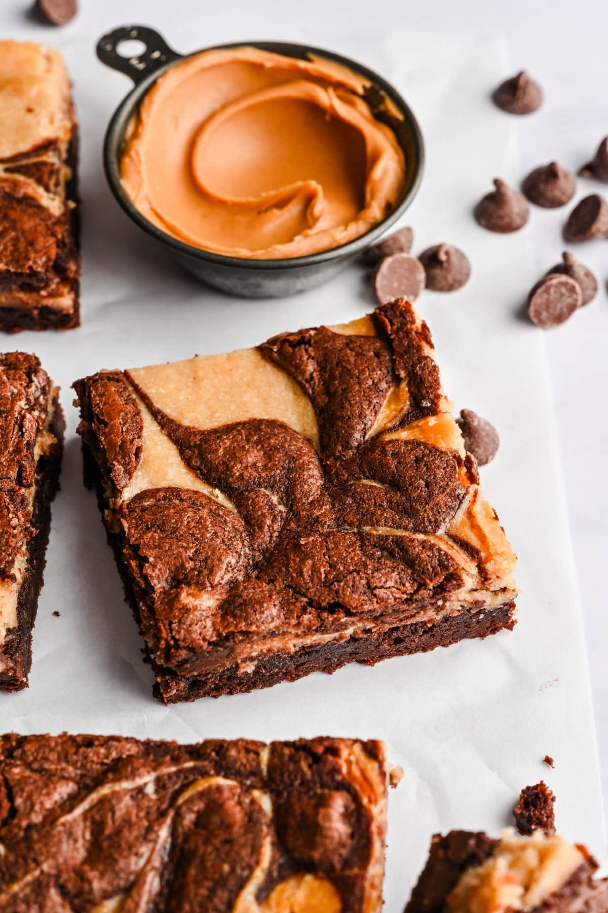 A peanut butter brownie next to a dish of peanut butter and scattered chocolate chips.