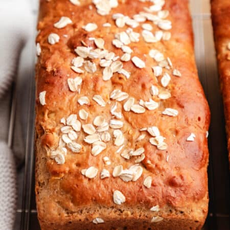 A loaf of honey oat bread on a wire cooling rack next to a knit dish cloth.