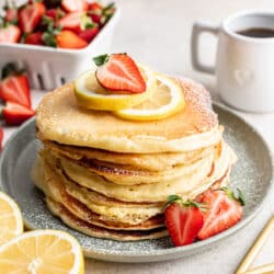 A stack of lemon ricotta pancakes topped with a slice of lemon and half a strawberry.