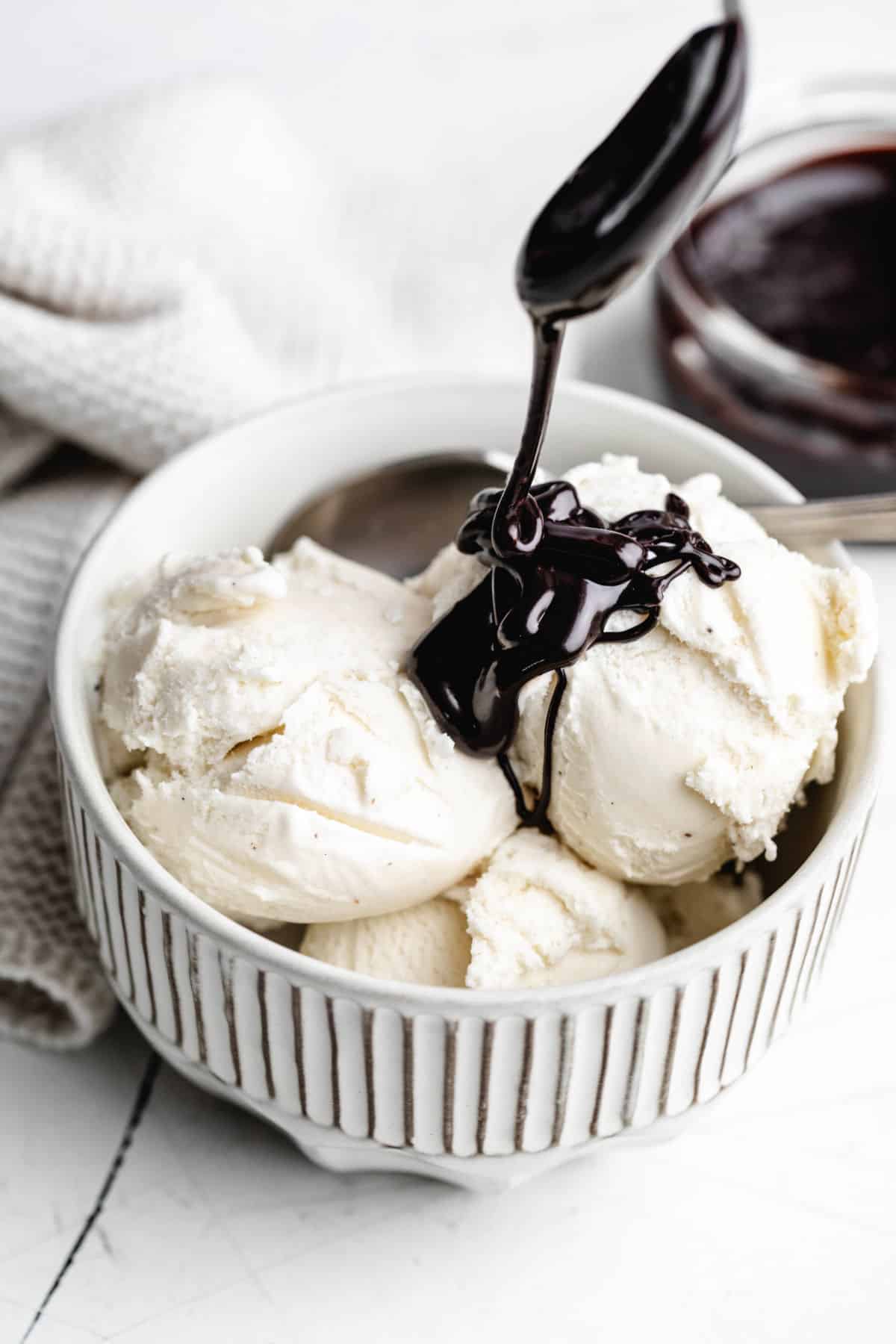 A spoon drizzling hot fudge over scoops of vanilla ice cream.