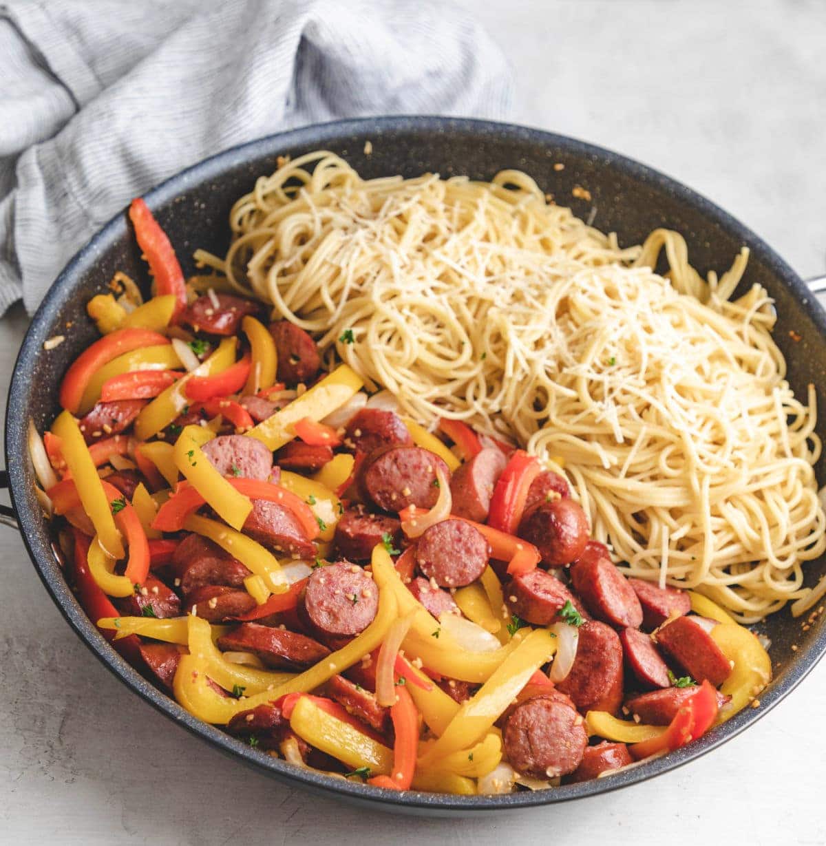 A skillet of peppers, onions, and kielbasa next to spaghetti in a black skillet.