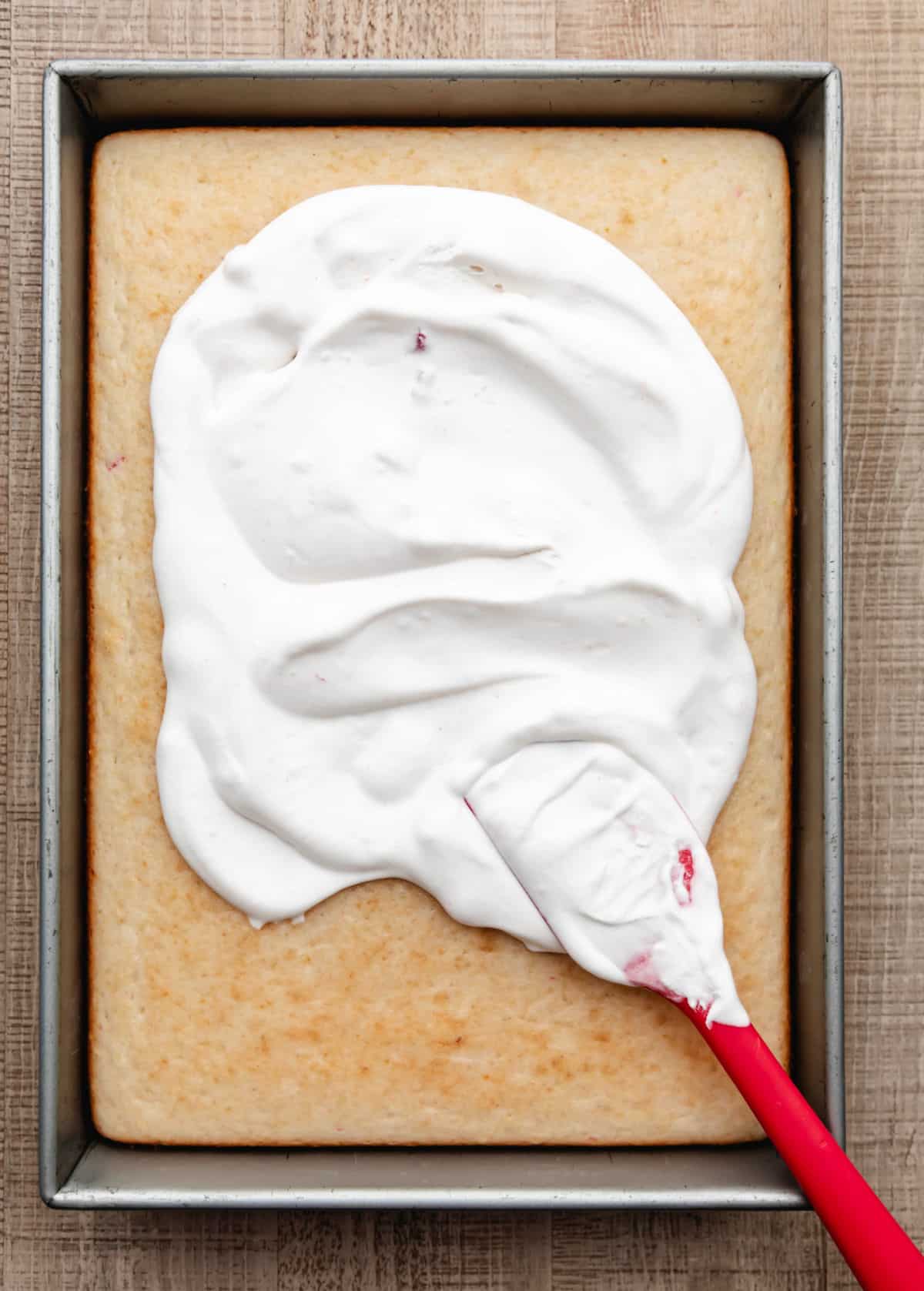 A red rubber spatula spreading frosting over a bake strawberry yogurt cake.