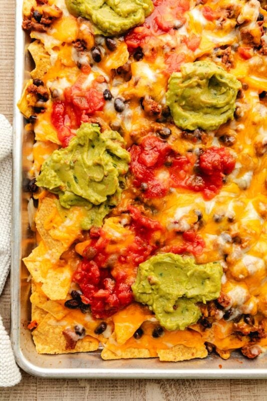 Sheet pan nachos in a rimmed baking sheet topped with guacamole and salsa.
