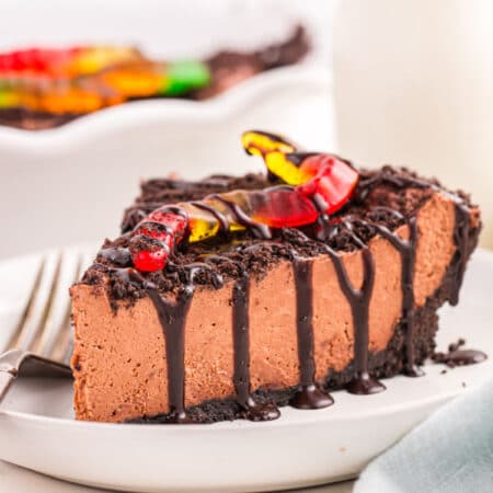 A slice of no bake dirt pie on a plate topped with chocolate syrup, crushed oreos, and a gummy worm.