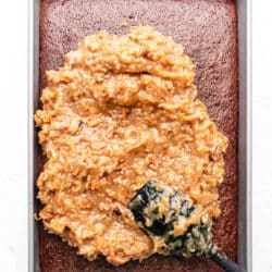 A black spatula spreading coconut pecan frosting over a chocolate cake.