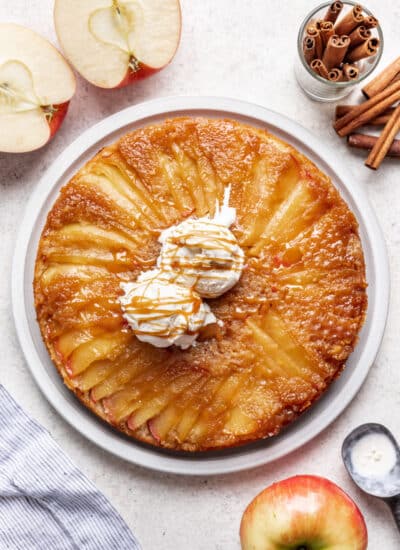 An apple upside down cake next to apples and cinnamon sticks.