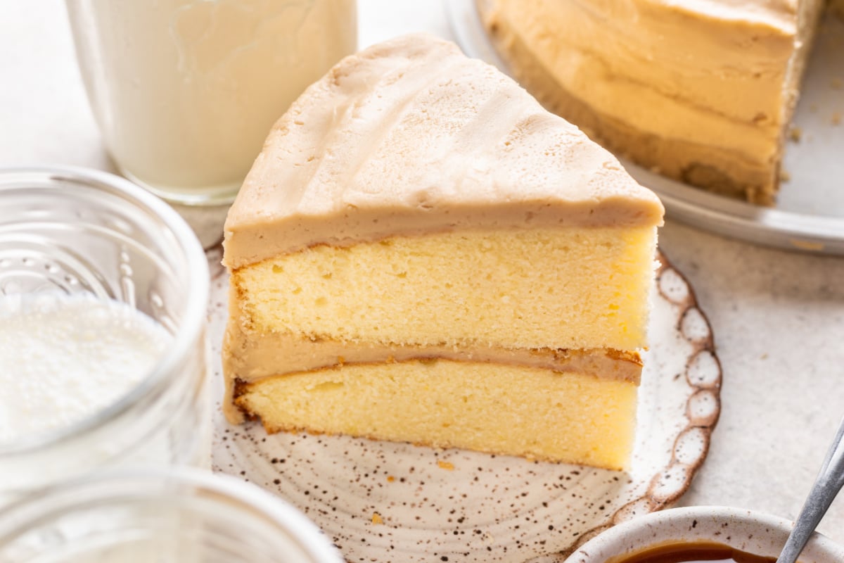 A slice of caramel cake on a plate next to a cup of milk.