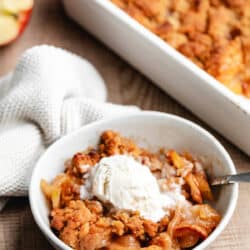 A bowl of apple crumble topped with vanilla ice cream next to the pan of crumble.