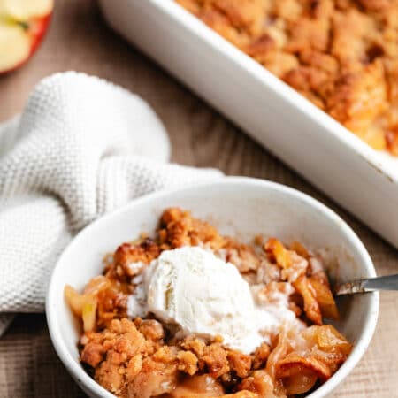 A bowl of apple crumble topped with vanilla ice cream next to the pan of crumble.