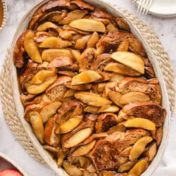 An oval baked filled with apple baked French toast next to a bowl of apples and syrup.