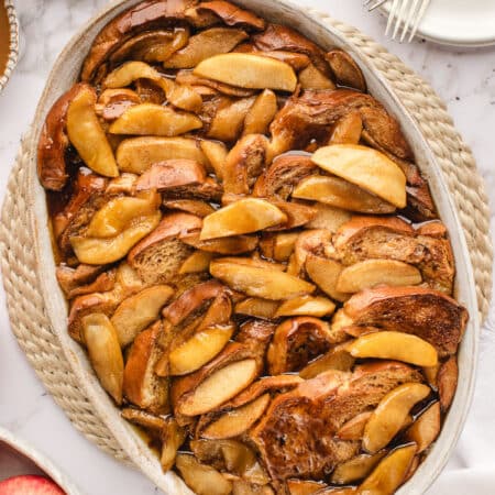 An oval baked filled with apple baked French toast next to a bowl of apples and syrup.