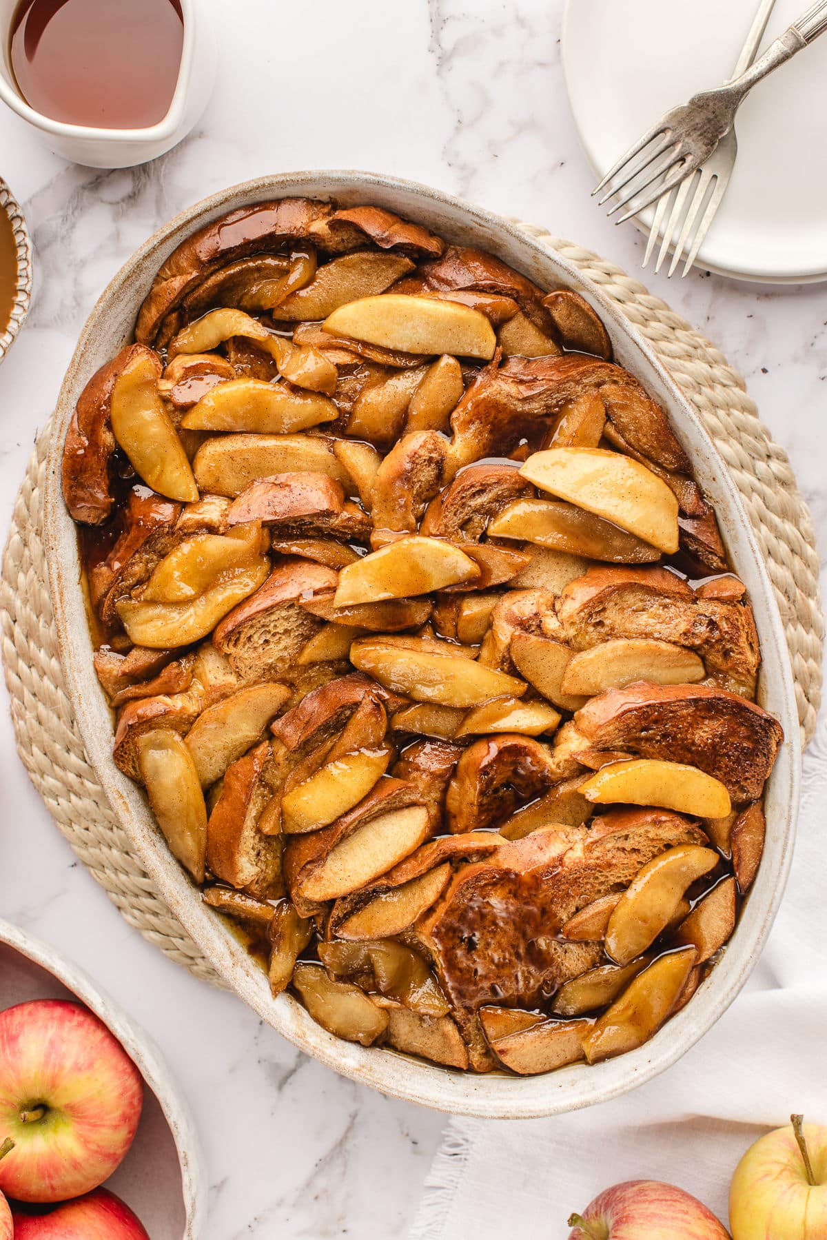 An oval baked filled with apple baked French toast next to a bowl of apples and syrup.