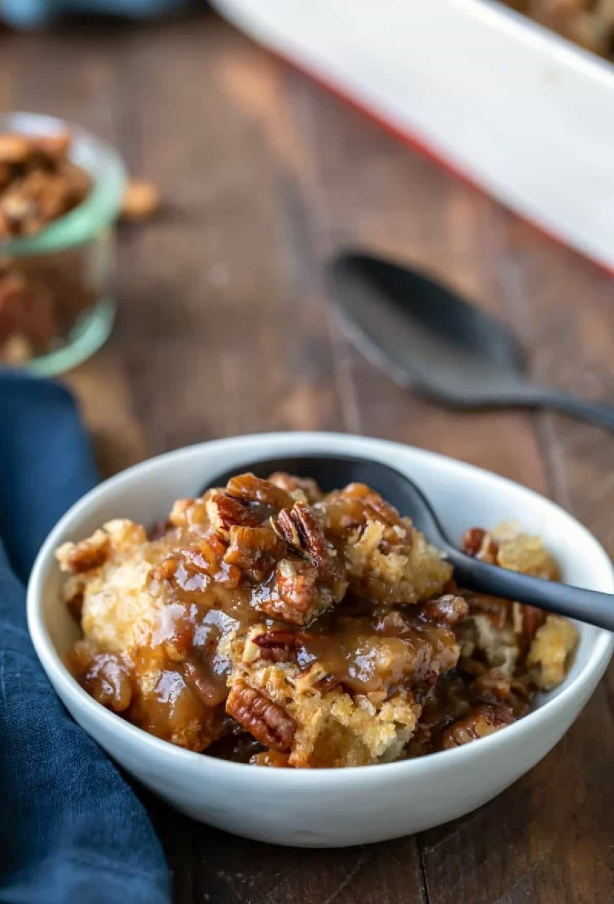 A white dish of pecan pie pudding cake with a black spoon in it.