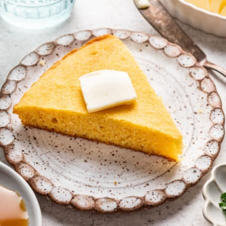A slice of cornbread with a pat of butter in top next to a dish of honey.