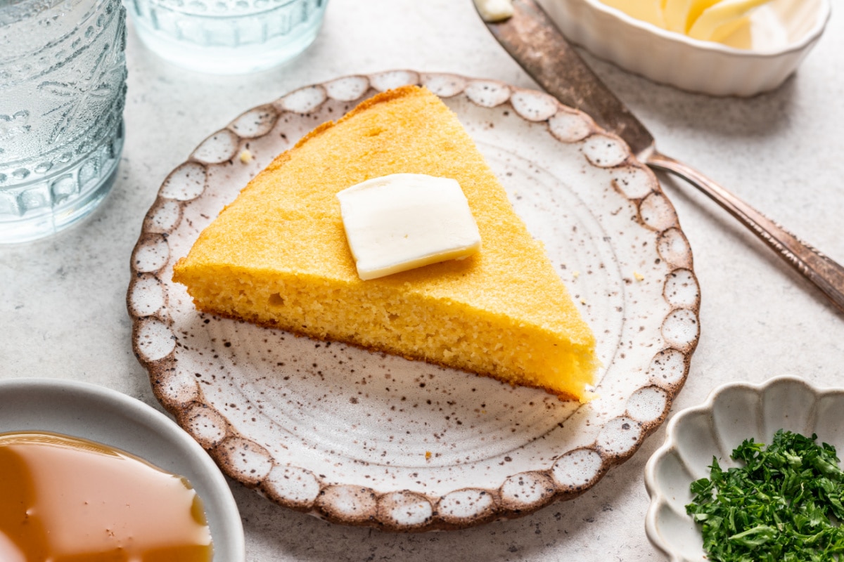 A slice of cornbread with a pat of butter in top next to a dish of honey.