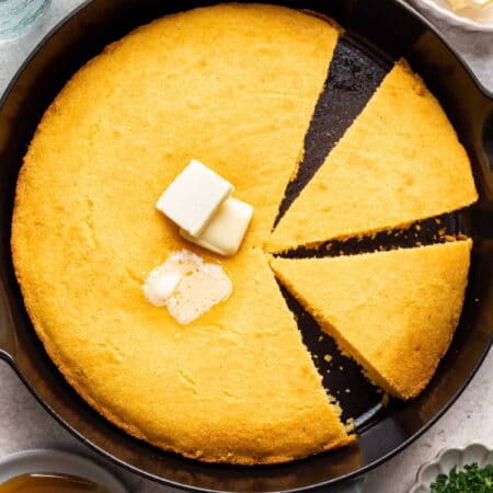 A skillet of cornbread with pats of butter in the middle with two pieces cut.