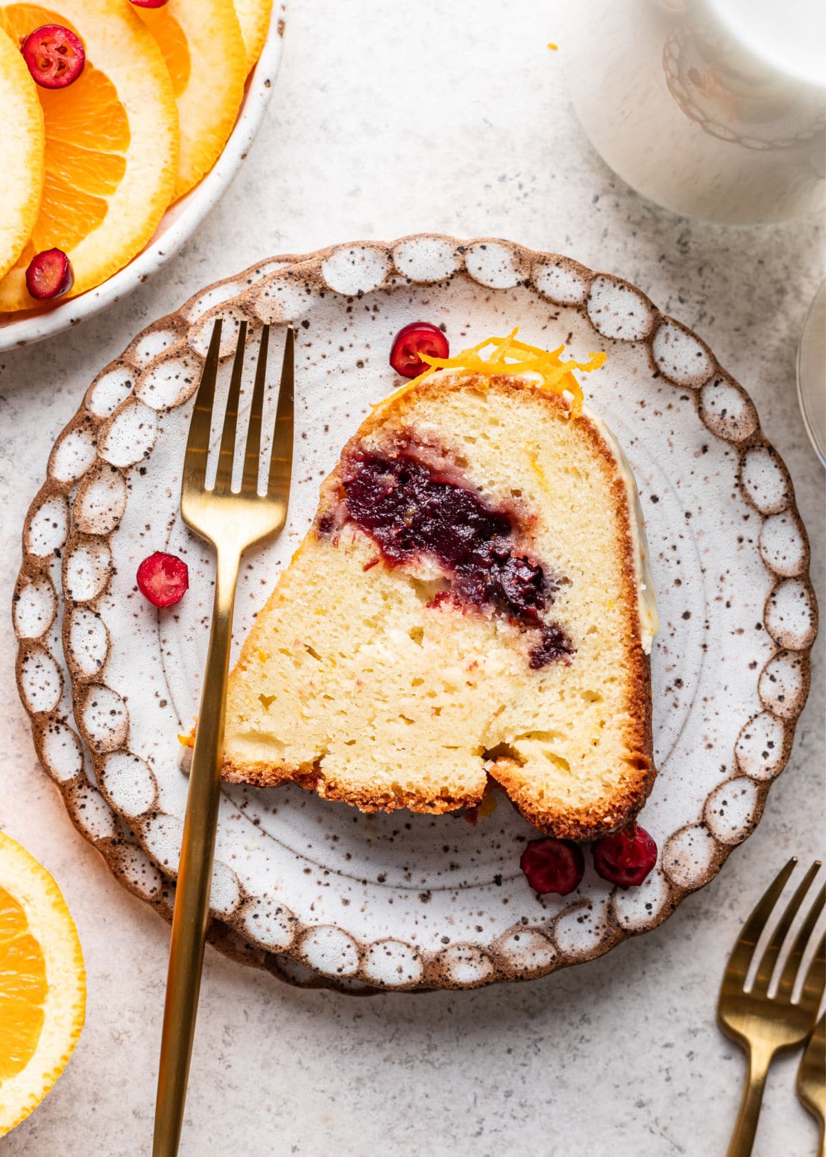 A slice of cranberry orange pound cake and a gold fork on a plate next to slices of fresh orange and cranberries.