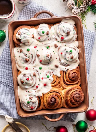 A pan of partially frosted cinnamon rolls next to cups of coffee and a dish of frosting.