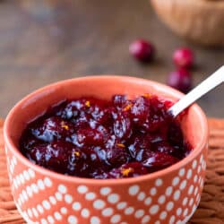 An orange dish of maple cranberry sauce next to a dish of fresh cranberries.
