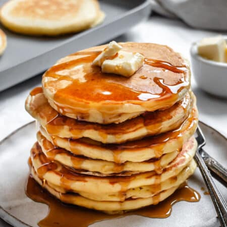 A stack of sour cream pancakes on a white plate next to a griddle.