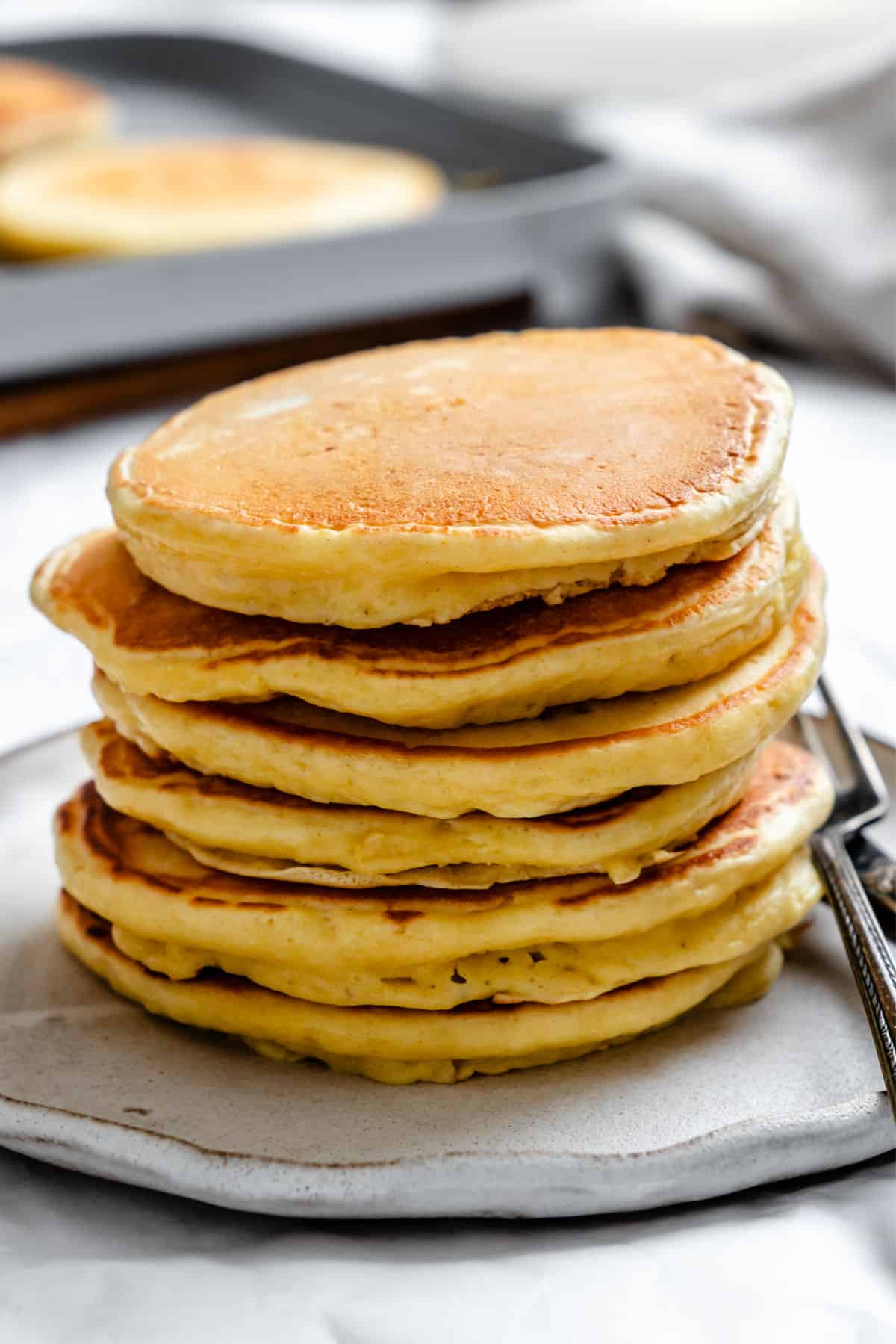 A stack of sour cream pancakes on a white plate with a silver fork next to them.