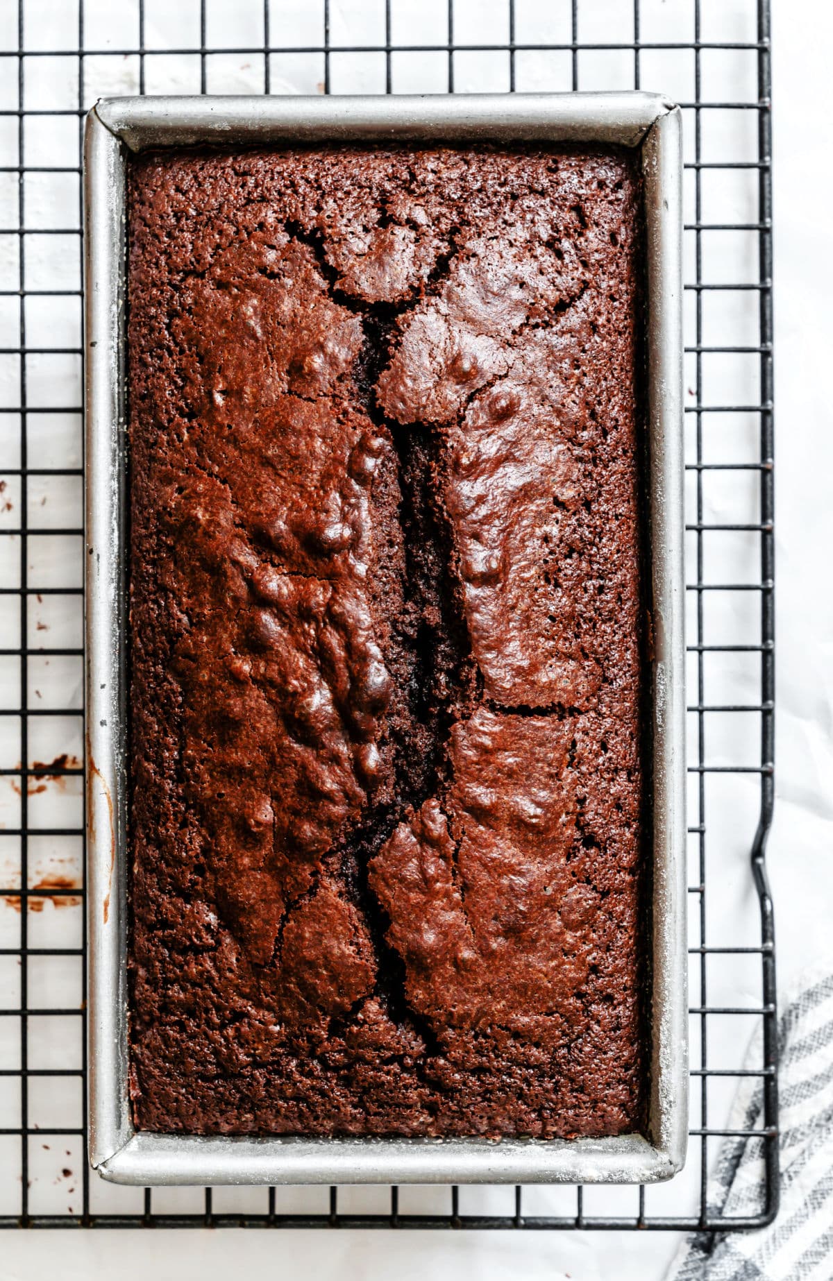 A loaf of chocolate pound cake in a loaf pan on a wire cooling rack.