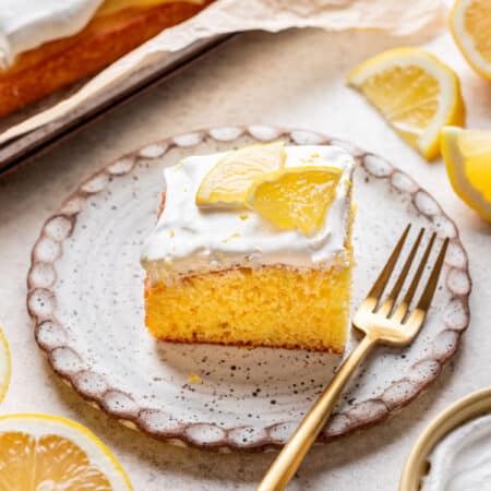 A piece of lemon poke cake on a plate with a gold fork next to it.