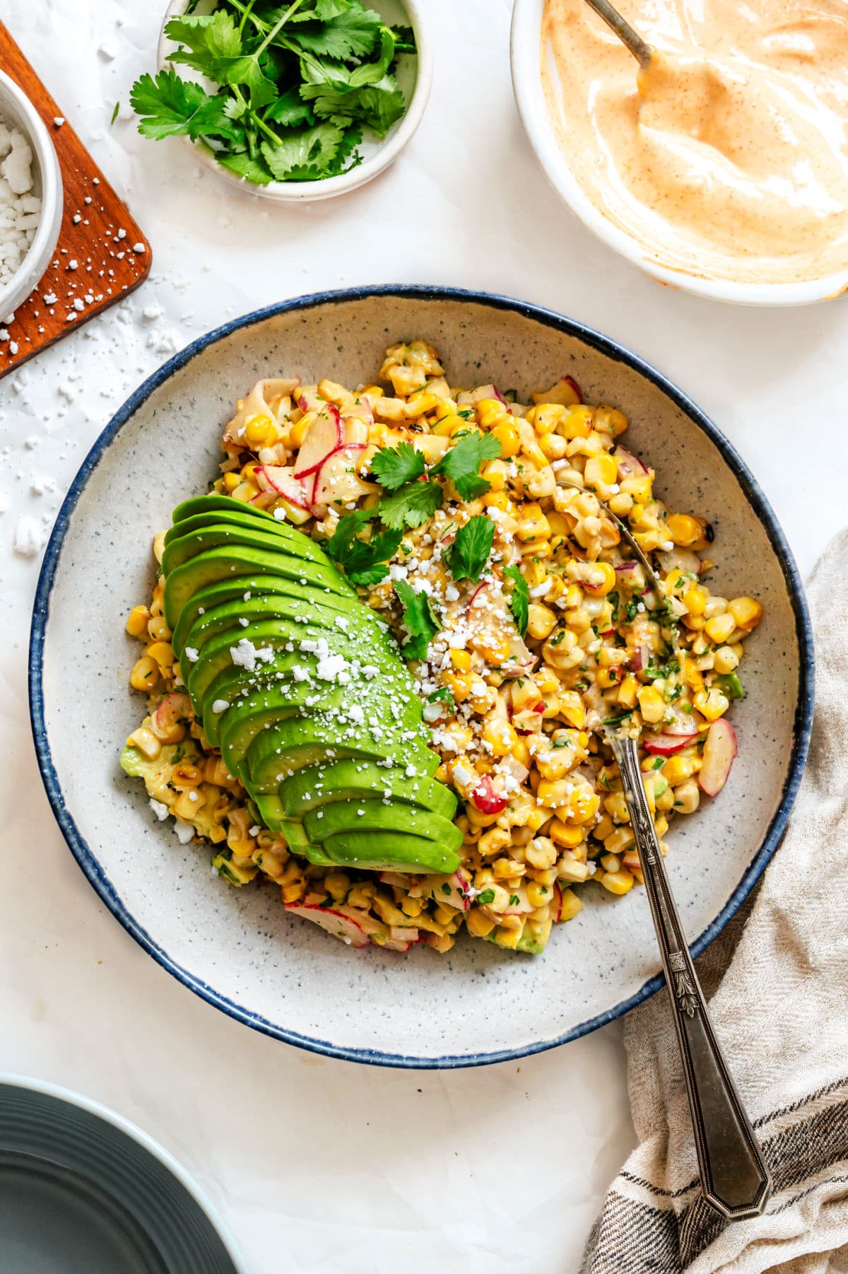 A bowl of Mexican street corn salad topped with sliced avocado and cotija cheese.