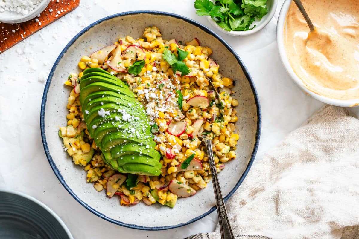 A dish of Mexican street corn salad next to dishes of chipotle dressing and fresh cilantro.