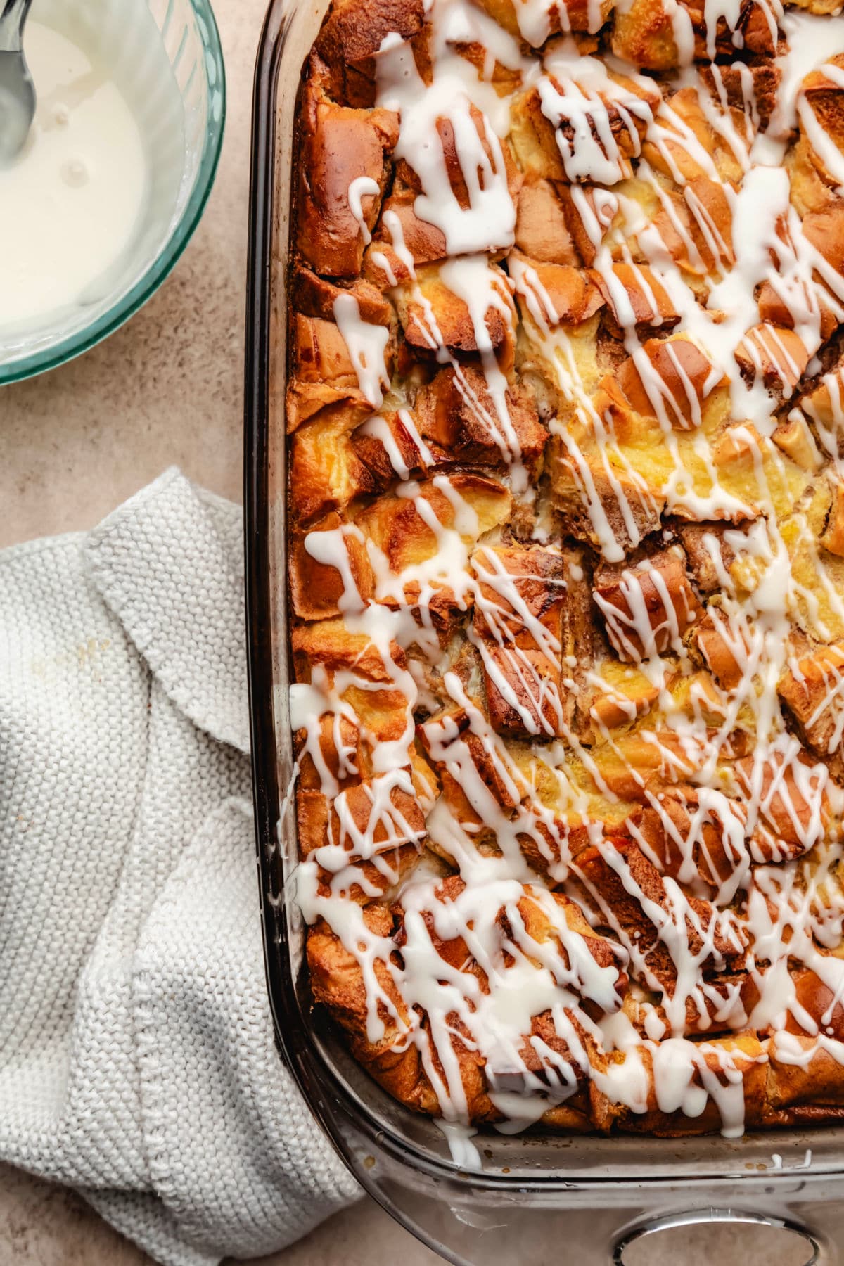 A baking dish of cinnamon roll bread pudding next to a bowl of icing. 