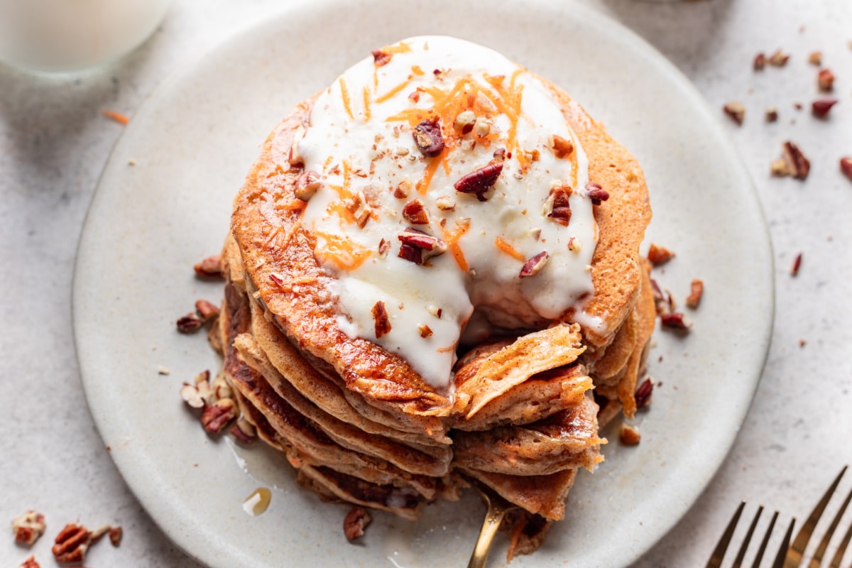 A fork holding a bite of carrot cake pancakes next to a stack of pancakes.