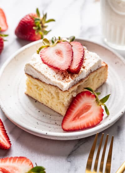 A slice of tres leches cake on a plate next to half of a sliced strawberry.