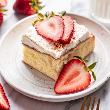 A slice of tres leches cake on a plate next to half of a sliced strawberry.