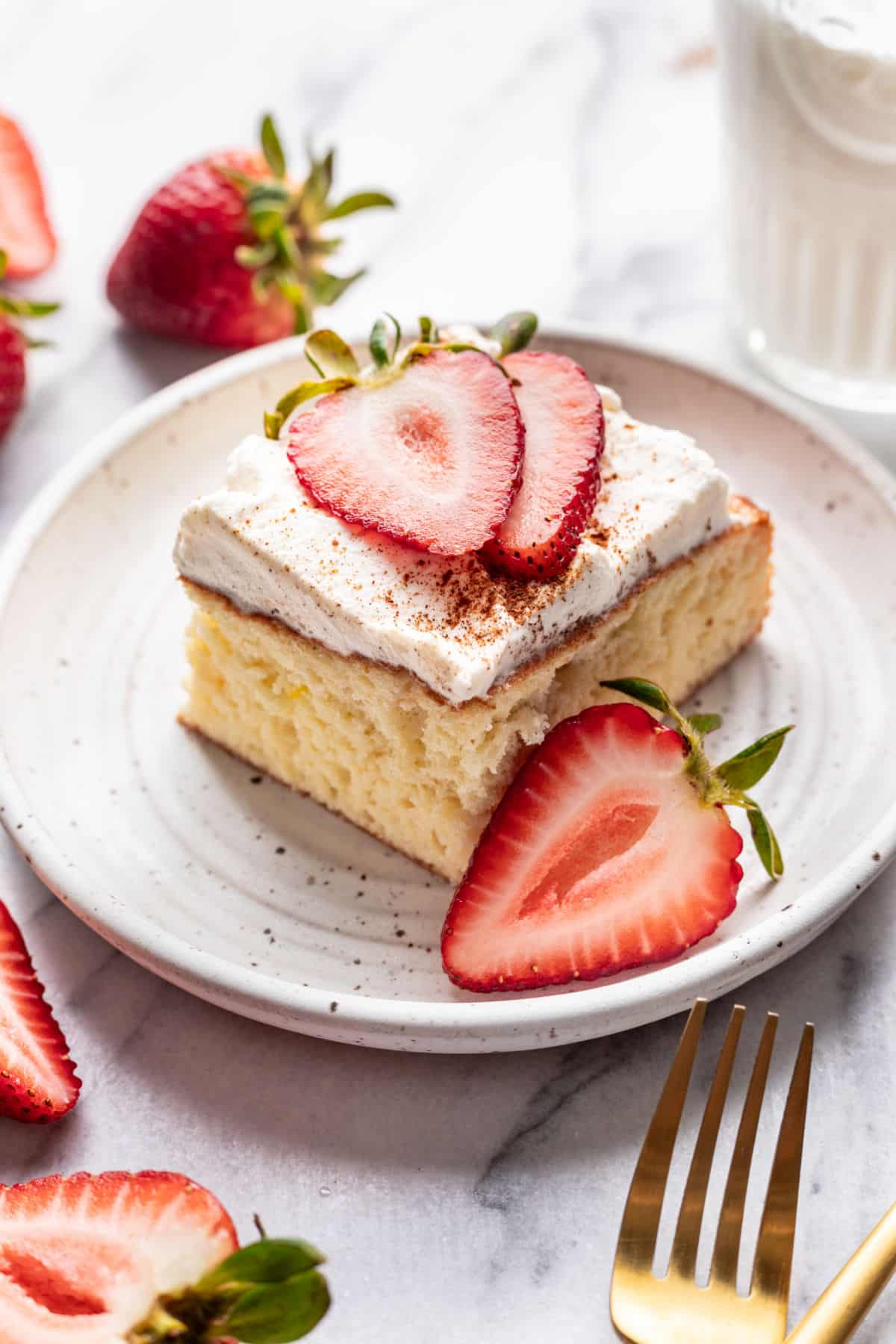 A slice of tres leches cake on a plate next to half of a sliced strawberry.
