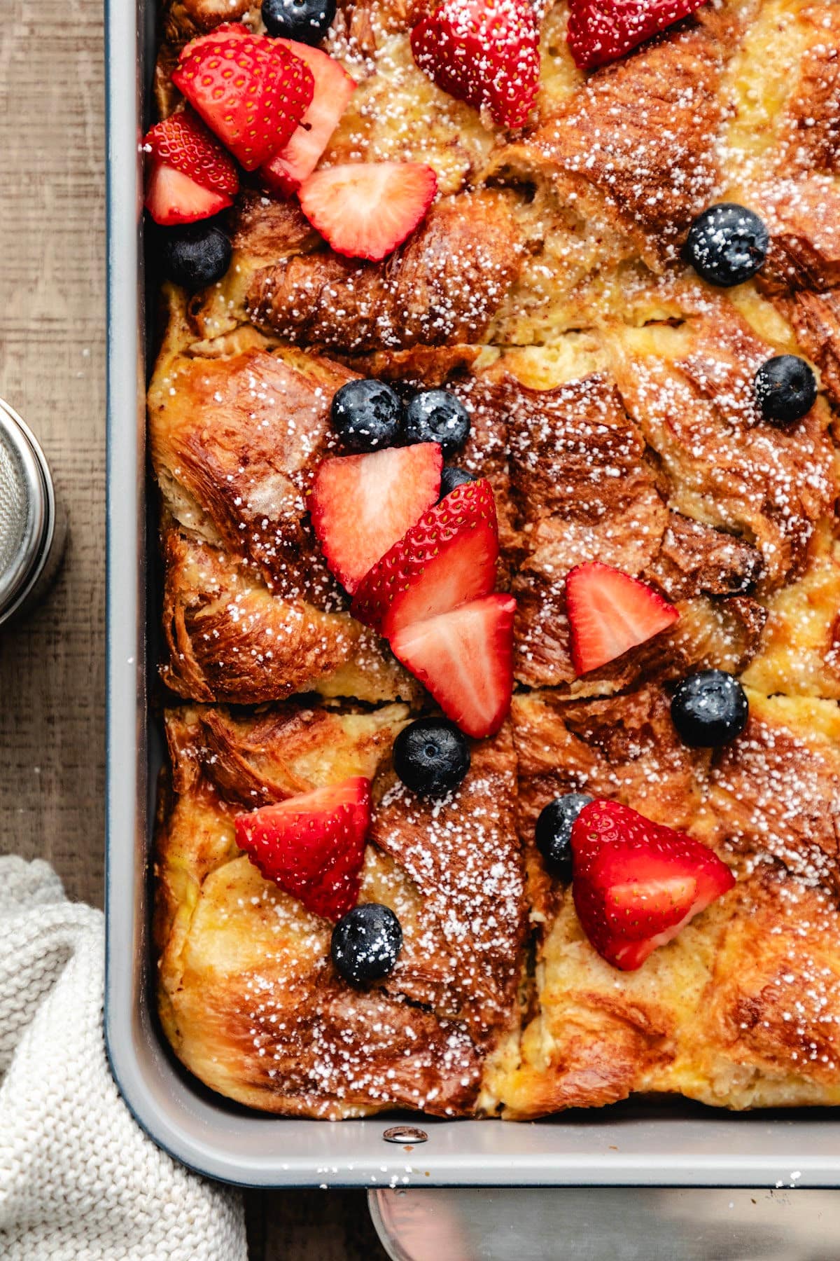 Close up photo of a pan of croissant baked French toast topped with powdered sugar and berries.