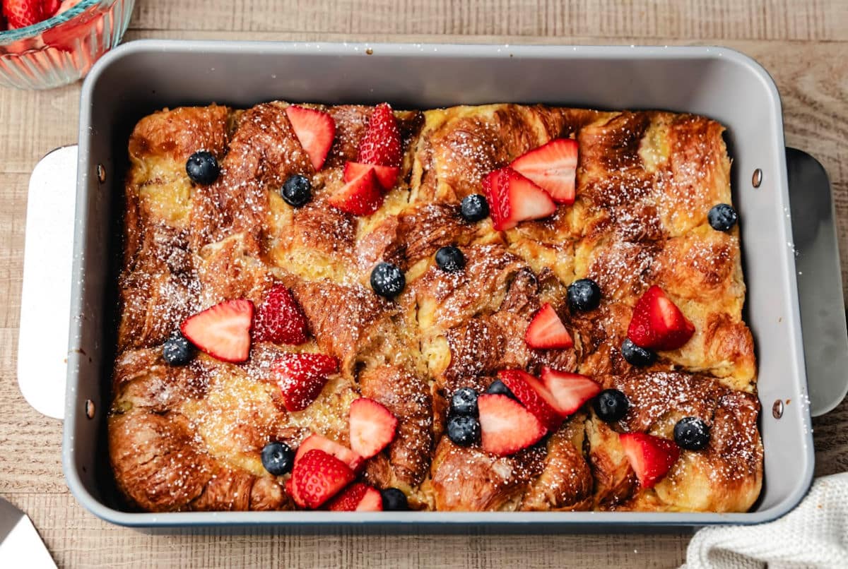 A pan of croissant baked French toast next to a bowl of cut strawberries.