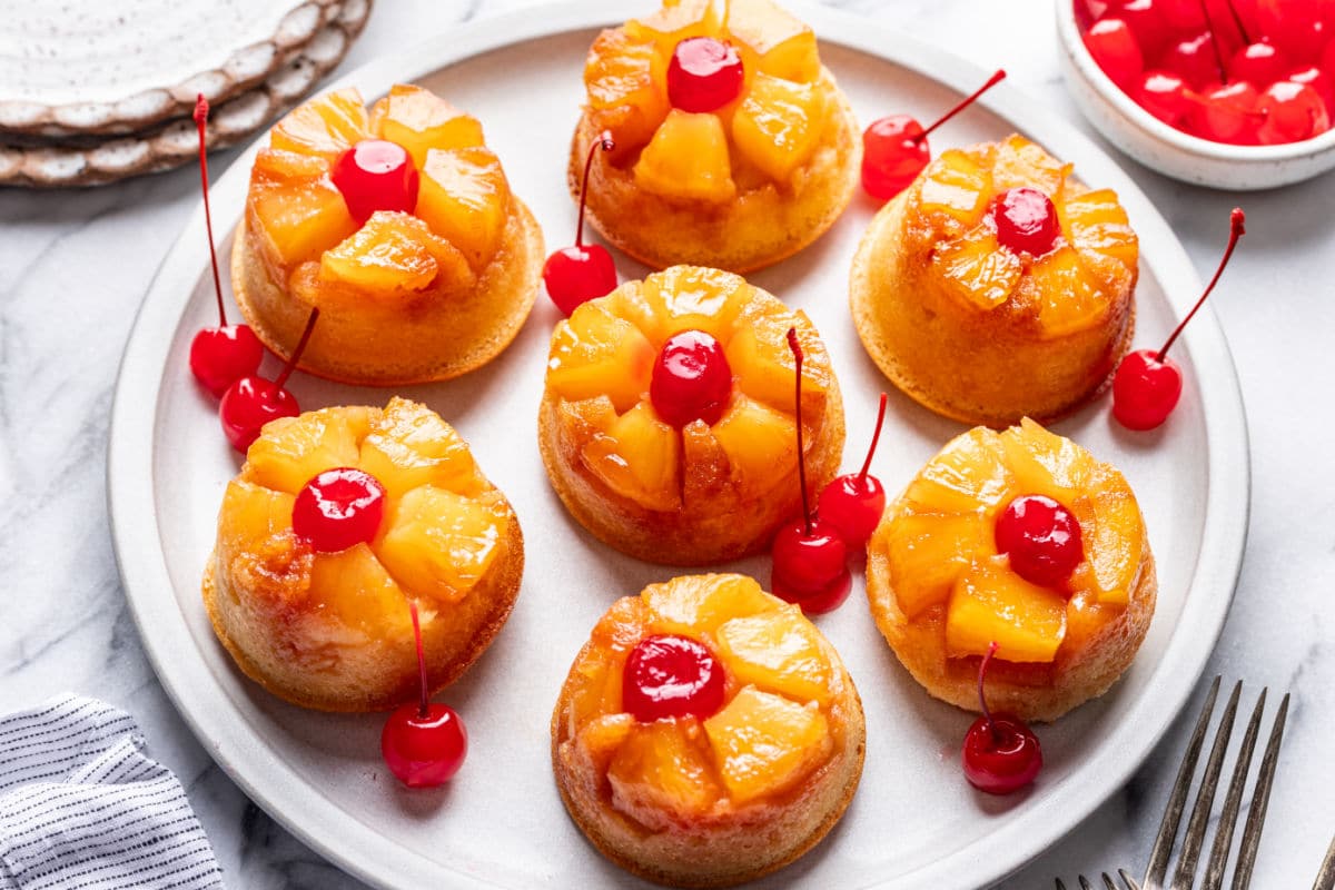 Pineapple upside down cupcakes on a platter next to a dish of maraschino cherries.