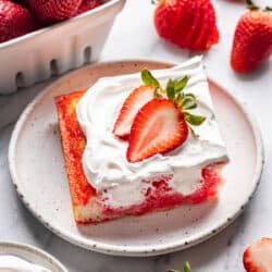 A slice of Jello Poke cake on a plate surrounded by fresh strawberries.