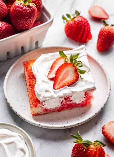 A slice of Jello Poke cake on a plate surrounded by fresh strawberries.