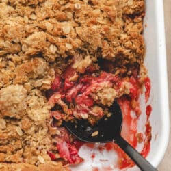 A black spoon holding a scoop of strawberry crisp in a white pan.