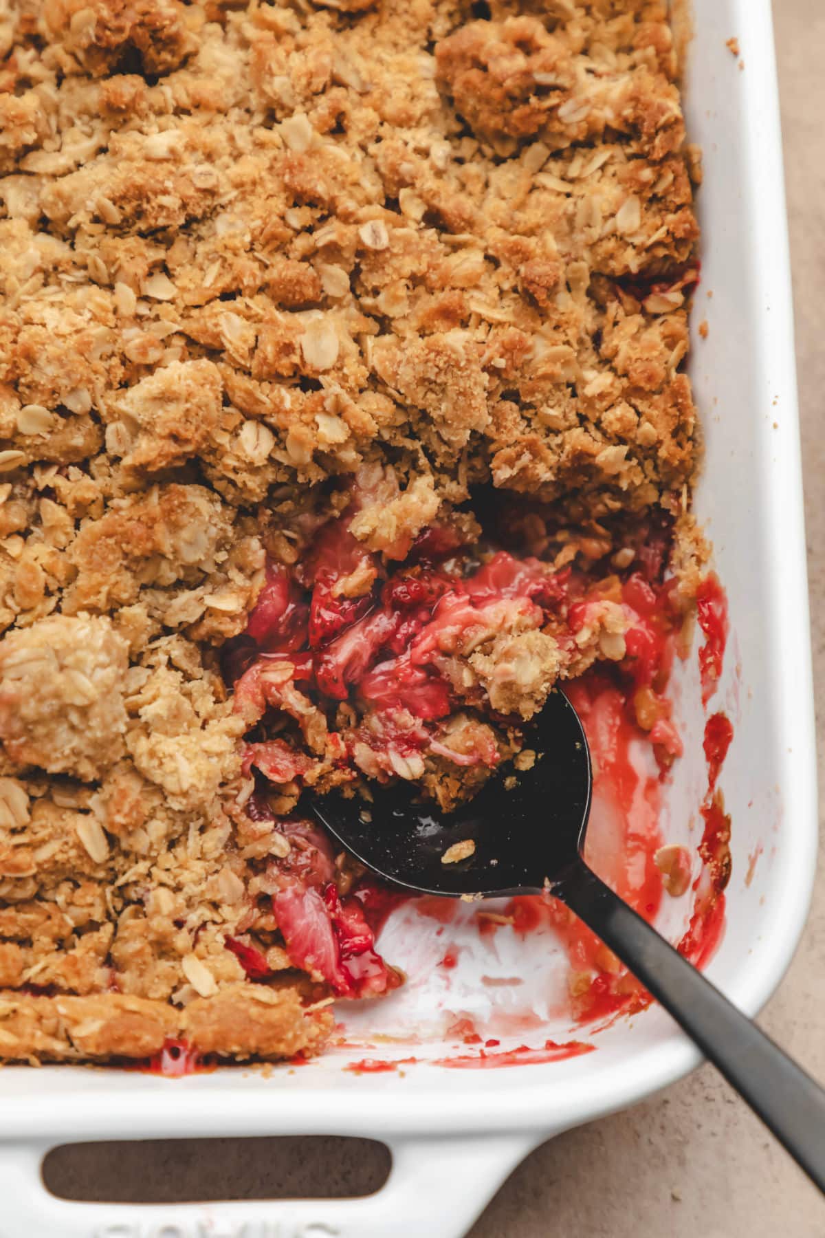 A black spoon holding a scoop of strawberry crisp in a white pan.