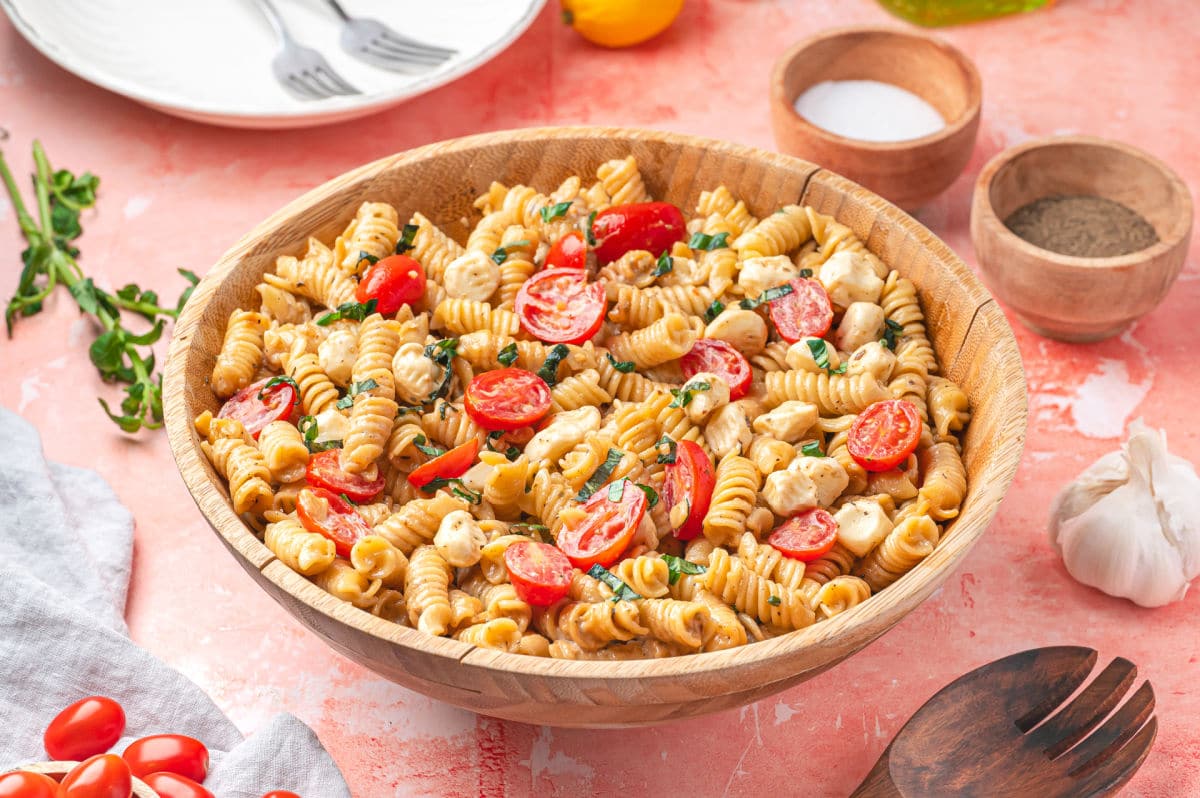 Un bol de salade de pâtes caprese à côté de plats de sel et de poivre et de tomates fraîches.