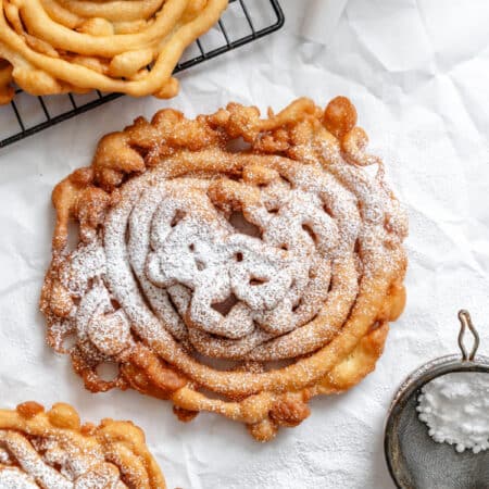 A funnel cake topped with powdered sugar next to a powdered sugar sifter.