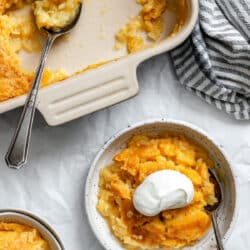 A bowl of pineapple dump cake topped with whipped cream next to the cake baking pan.
