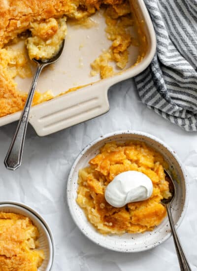 A bowl of pineapple dump cake topped with whipped cream next to the cake baking pan.